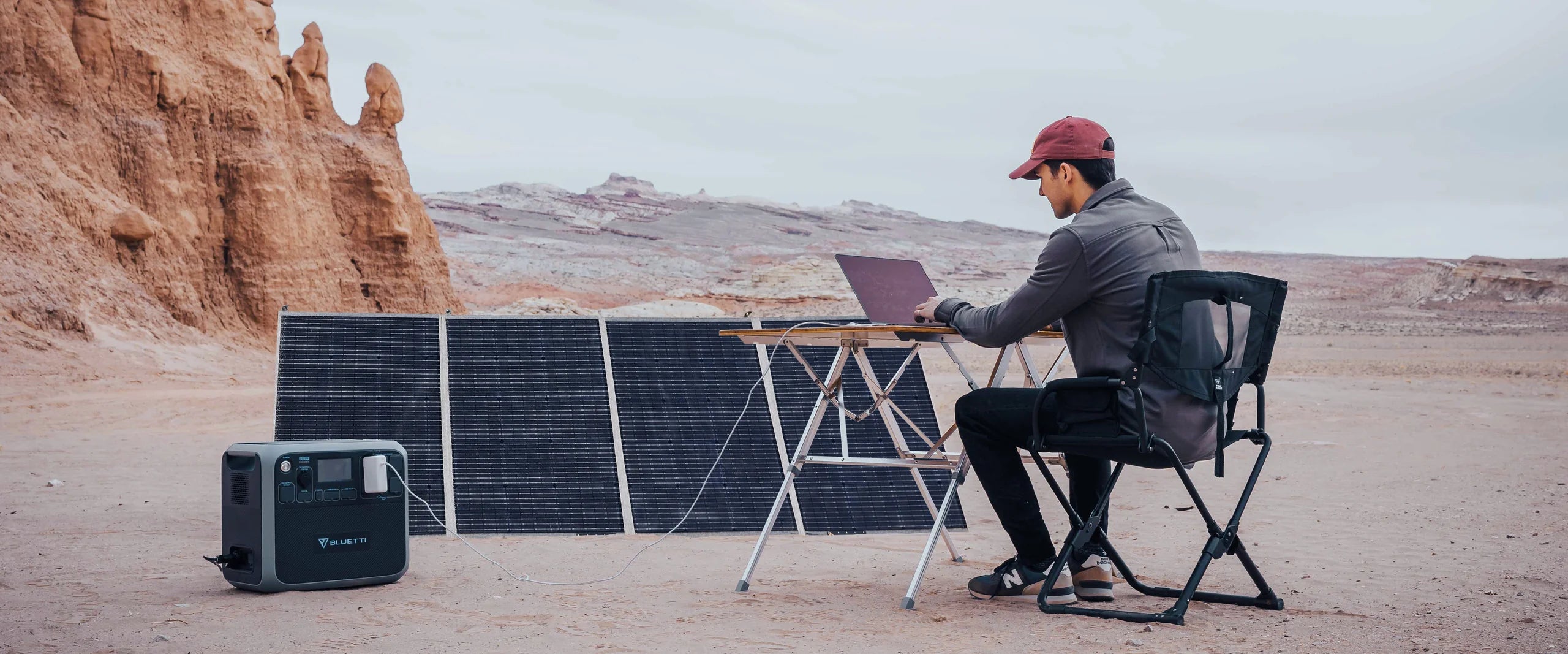 A person works on a laptop at a portable desk in a rocky desert, powered by the BLUETTI 420W 48V Monocrystalline Portable Solar Panel from Bluetti, which reliably collects solar energy for their portable station.