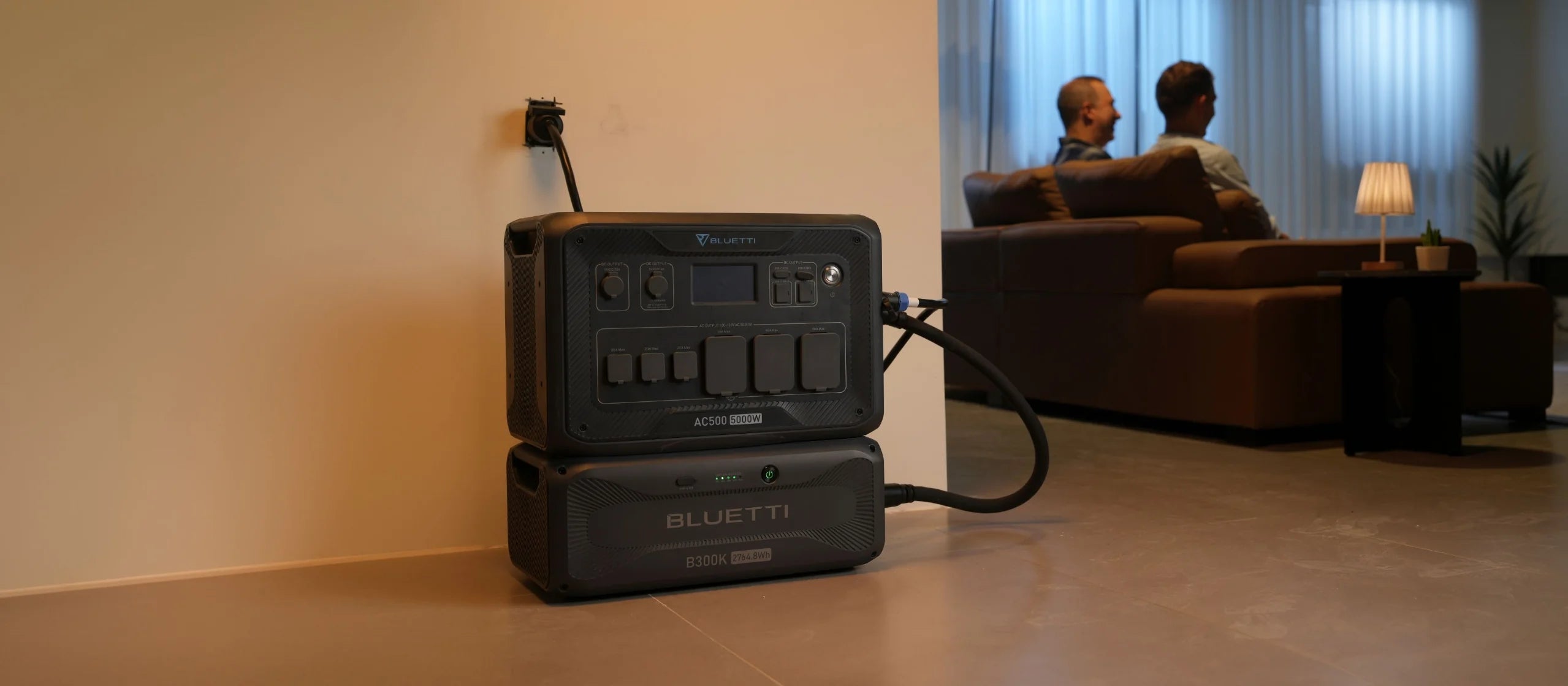 A Bluetti AC500+B300K 5,000W solar generator kit powers a cozy living room, where two people relax on a sofa with warm lighting and a side table lamp. The complete solar kit includes 3 x 200W mono solar panels.