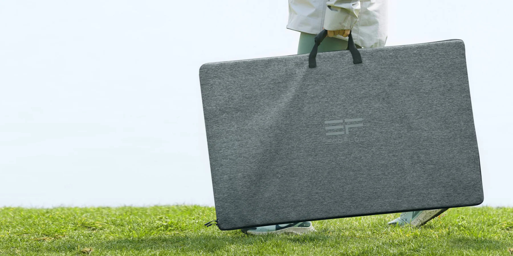 EcoFlow 400W 48V Monocrystalline Portable Solar Panel A person stands on grass holding the EcoFlow 400W 48V Monocrystalline Portable Solar Panel case, which is large, gray, branded with the EcoFlow logo, and foldable—nearly covering their legs except for their lower legs and shoes.