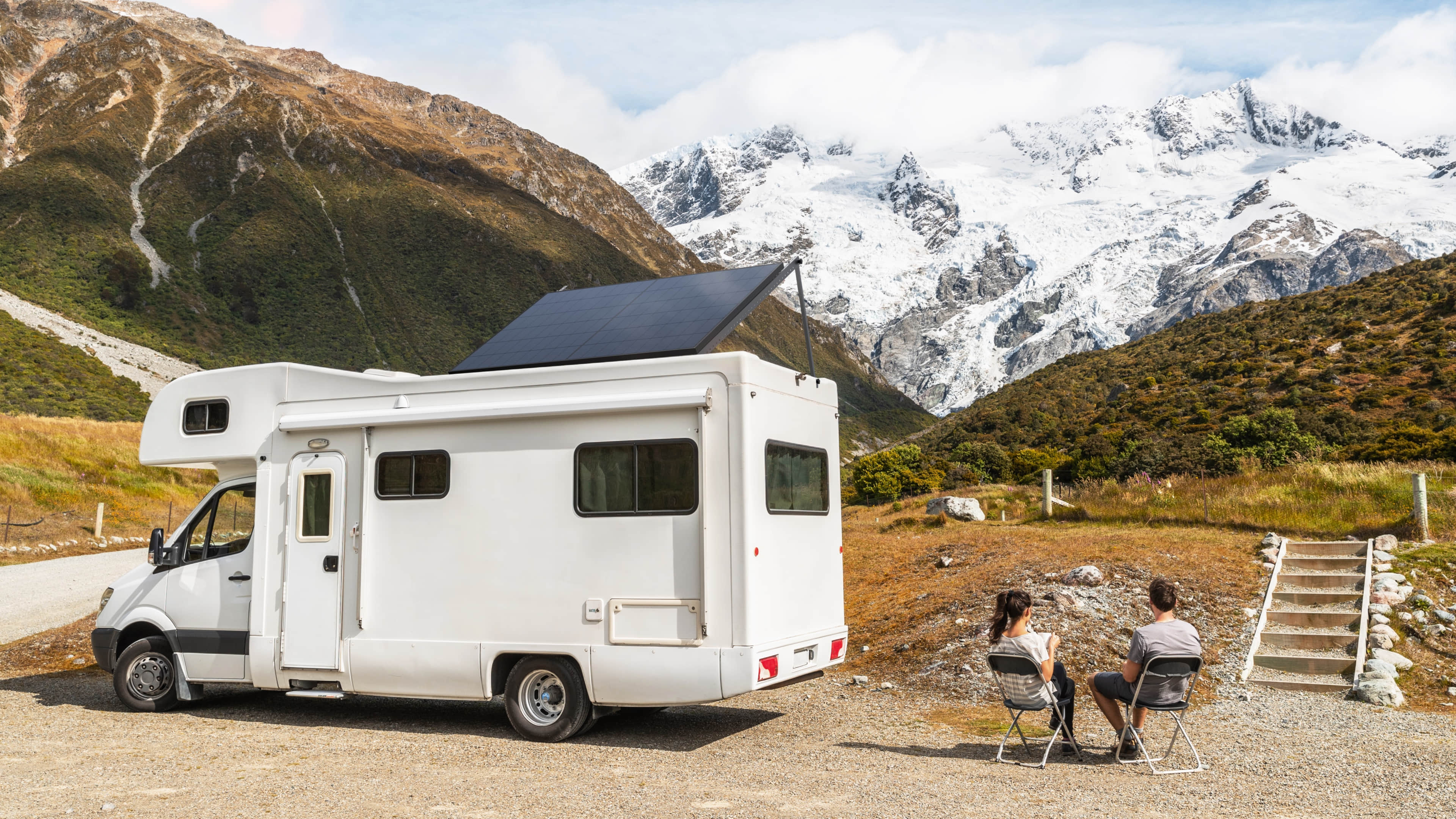 EcoFlow 400W 24V Monocrystalline Rigid Solar Panel A white RV with an EcoFlow 400W 24V Monocrystalline Rigid Solar Panel is parked by a gravel road; two people relax on folding chairs nearby, enjoying views of snow-capped mountains and green hills beneath a partly cloudy sky.