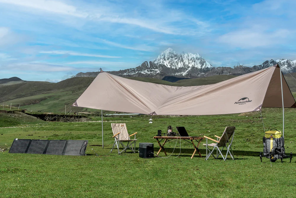 A grass campsite includes two chairs, a table with gear, a large tan canopy, an EcoFlow 110W 24V Monocrystalline Portable Solar Panel by EcoFlow, a backpack, and snowy mountain peaks beneath a blue sky.