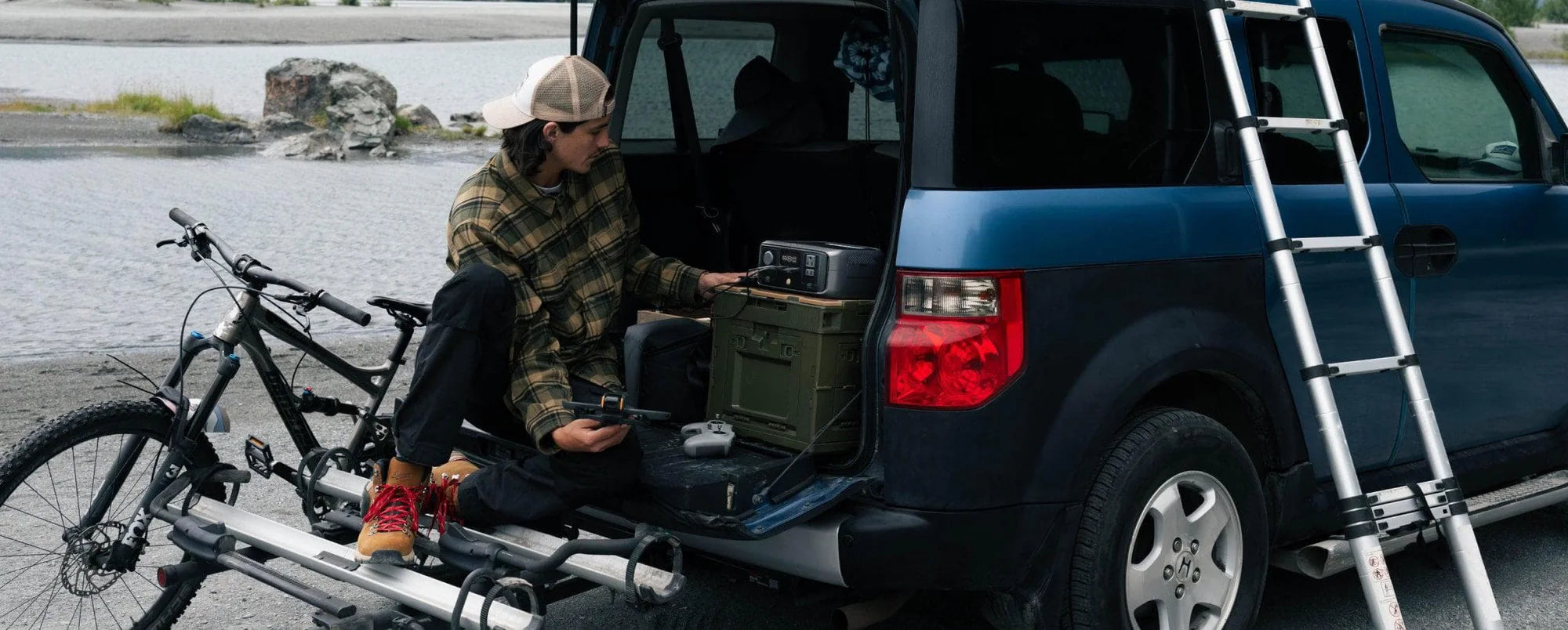 EcoFlow RIVER 3 300W 245Wh Portable Solar Power Station Wearing a plaid shirt and cap, someone sits on an SUV’s open tailgate by a lake, using the EcoFlow RIVER 3 300W 245Wh Portable Solar Power Station. A bicycle is mounted on the back and a ladder leans against the vehicle.