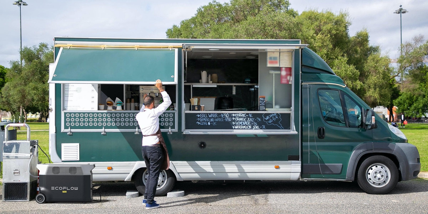 EcoFlow DELTA Pro 3600W 3.6kWh Portable Power Station + 400 Watts of Portable Solar Panels A person in an apron stands by a green food truck in a park, powered by the Ecoflow Delta Pro 7,200W Battery Backup with 6 x 200W Mono Solar Panels, raising their arm near the serving window with menu and equipment visible.