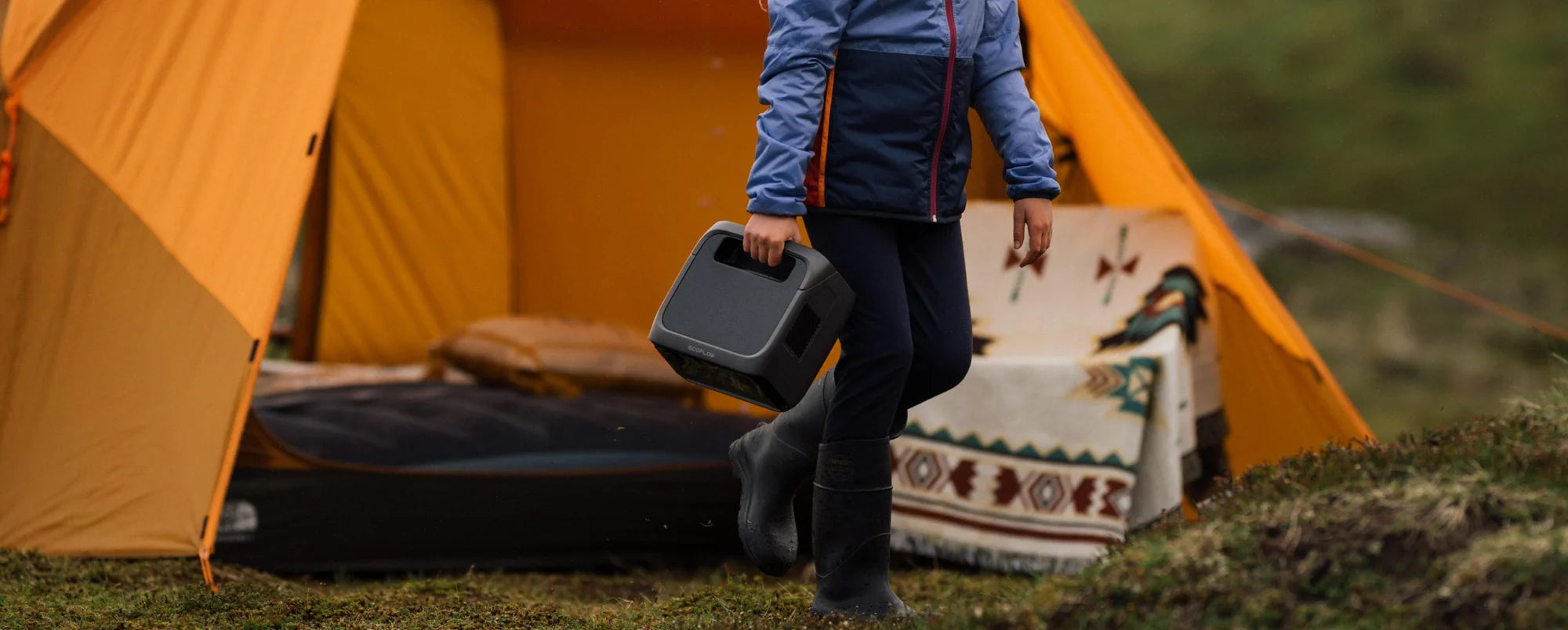 Ecoflow RIVER 3 Max 600W 572Wh Portable Solar Power Station | River 3 Plus + RIVER 3 Plus Smart Extra Battery (286Wh) A person in a blue jacket and black boots carries an EcoFlow RIVER 3 Plus 600W 286Wh Portable Solar Power Station outside a yellow camping tent on grass, with a patterned blanket and air mattress inside the tent.