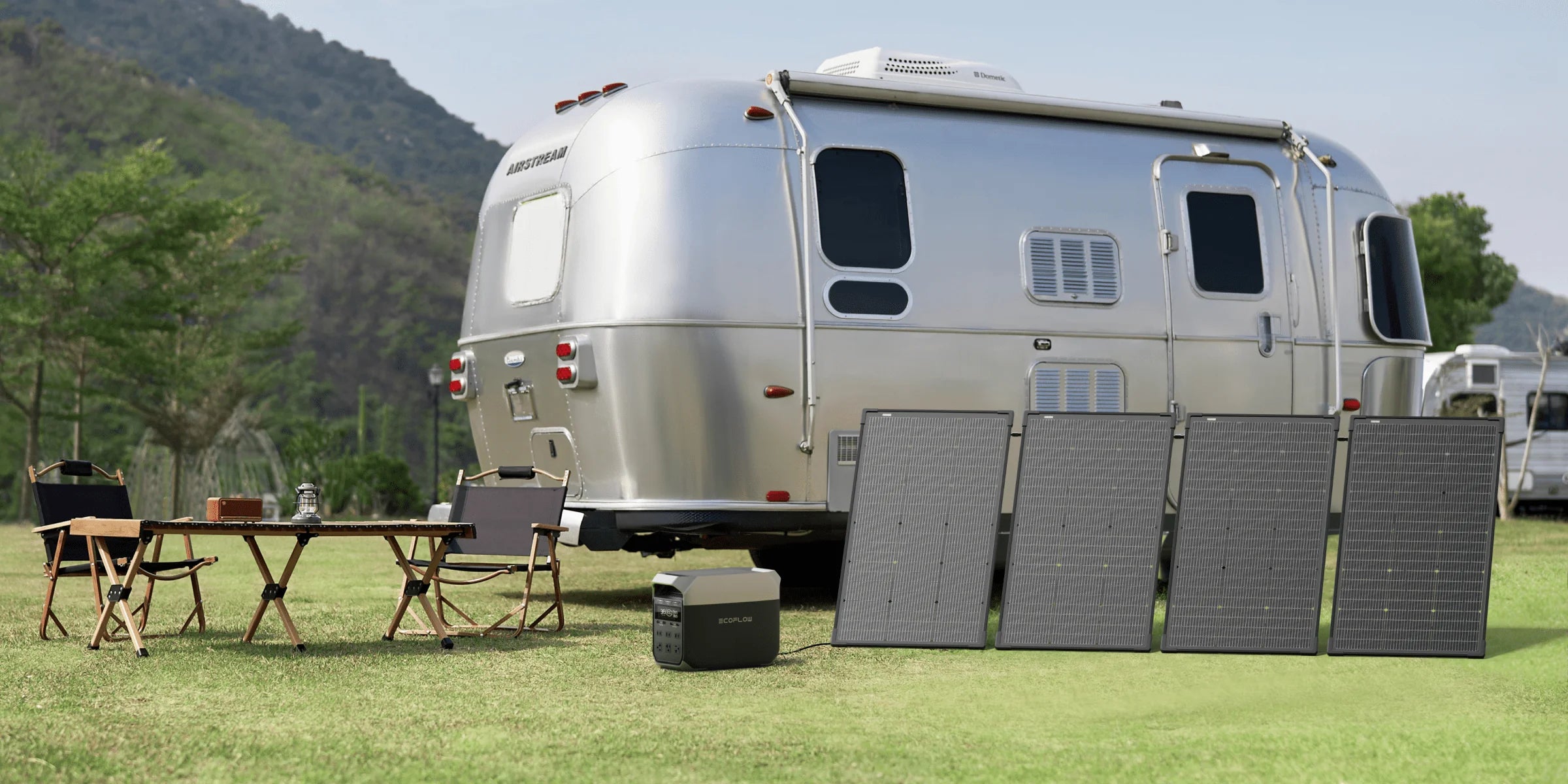 EcoFlow 125W 48V Monocrystalline Bifacial Modular Solar Panel A shiny silver camper trailer is parked on grass near mountains. In front, four Ecoflow 125W 48V Monocrystalline Bifacial Modular Solar Panels, a portable power station, a wooden table, and two folding chairs are set up for camping.