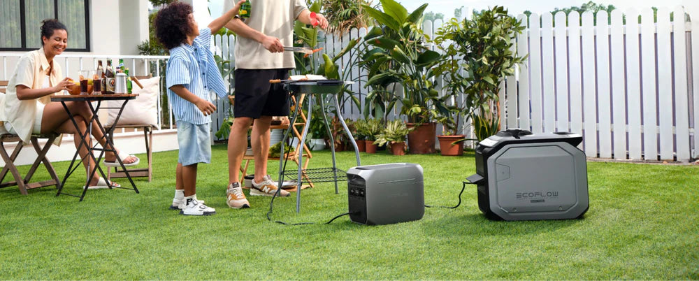 EcoFlow DELTA 3 Plus 1,800W 1,024Wh Plus Portable Solar Power Station A family enjoys a backyard barbecue as a woman sits at a table, while a man and child grill food powered by the EcoFlow DELTA 3 Plus 1,800W 1,024Wh Portable Solar Power Station. Potted plants and a white fence are in the background.