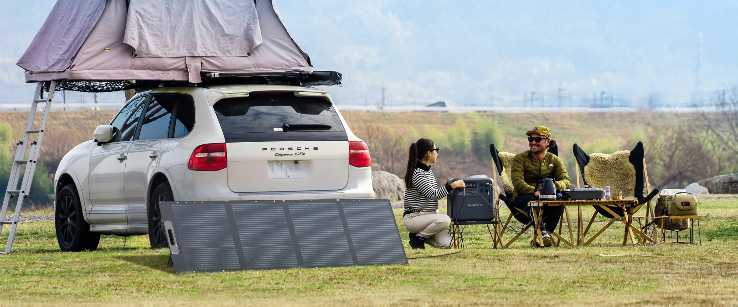 A white Porsche SUV with a rooftop tent is parked on grass. Two people sit beside it at a campsite with folding chairs, a table, and the Bluetti 200W 24V Monocrystalline Portable Solar Panel, enjoying an outdoor conversation.