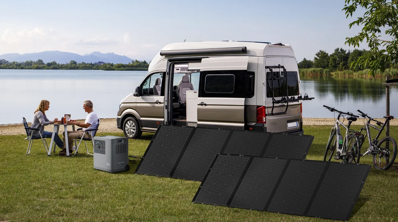 Mango Power E 3,000W 3.5kWh 120V Portable Power Station + 2 x 200W Solar Panels Package A couple sits at a table by a lake near their camper van, which is equipped with the Mango Power E 3,000W 3.5kWh 120V Portable Power Station and two 200W solar panels by Mango Power, along with two bicycles, as they enjoy an outdoor meal under a clear sky.
