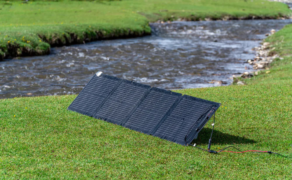 EcoFlow 110W 24V Monocrystalline Portable Solar Panel An EcoFlow 110W 24V Monocrystalline Portable Solar Panel by EcoFlow, lightweight and black, is set up on green grass near a river. Angled toward the sun, visible cables show its generating power outdoors.